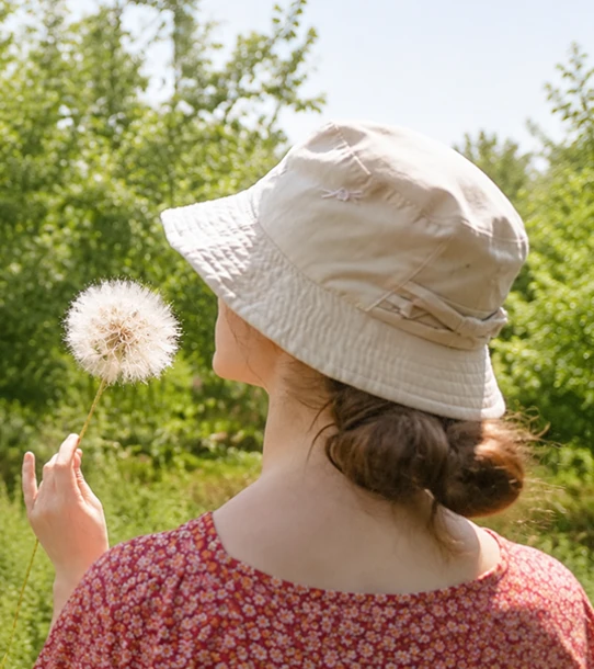 A woman in a sunhat holding a dandelion stands outdoors surrounded by greenery.